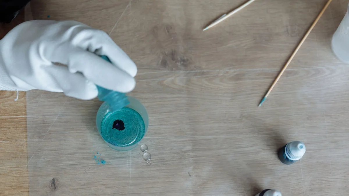 A gloved hand pouring blue resin into a container on a wooden table, creating art.