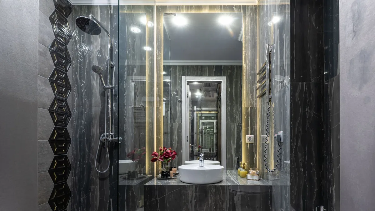 Interior of spacious contemporary bathroom with black tiled walls and floor with transparent shower cabin near bidet and sink at mirror